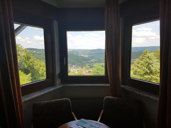 A room with a large window offering a view of a valley and hills. There are two chairs and a table with papers.