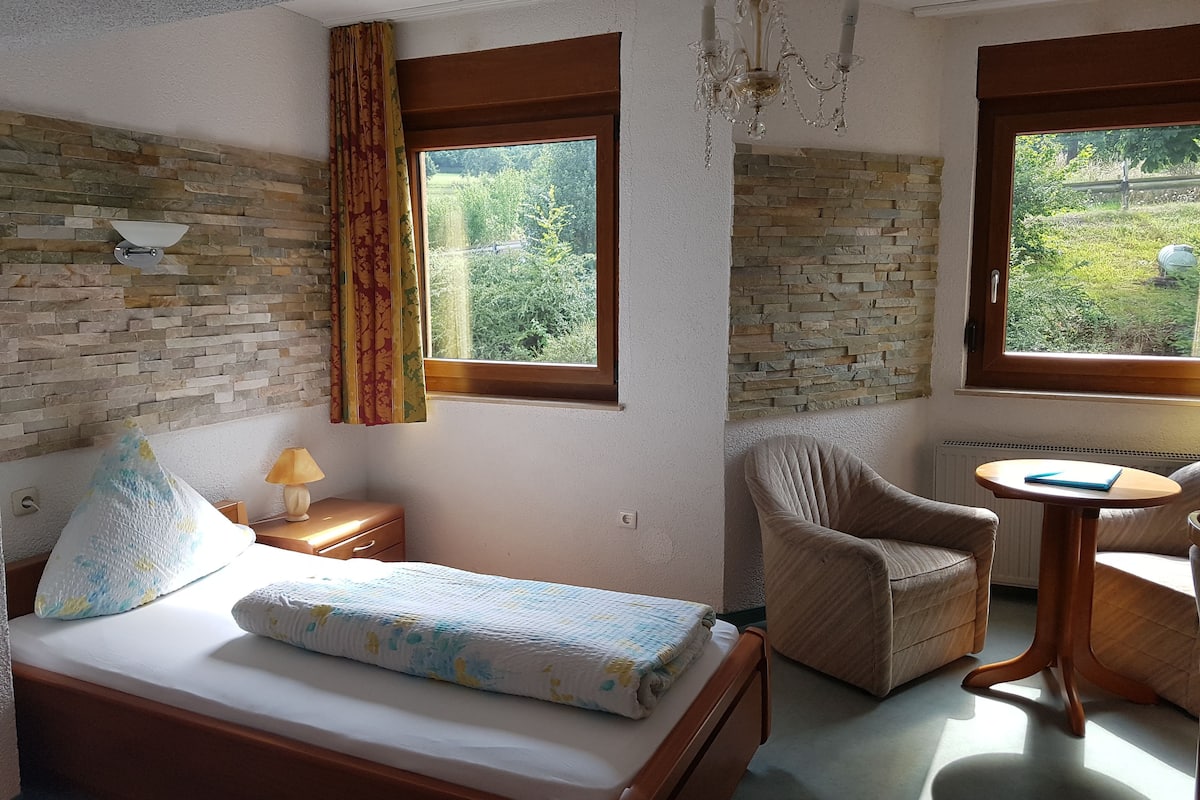 A bedroom with a bed, a chair, a table, a lamp, and a view of greenery through the windows.