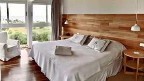 A bedroom with a large bed, two bedside tables, a chair, and a view of greenery outside. - Casa Bikini (Manatiales)