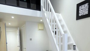 A modern living room with a dark grey sofa, a black and white patterned pillow, a wooden coffee table, and a staircase with white railings leading to an upper level. - 22 43 Clanricarde Gardens Studio (London)