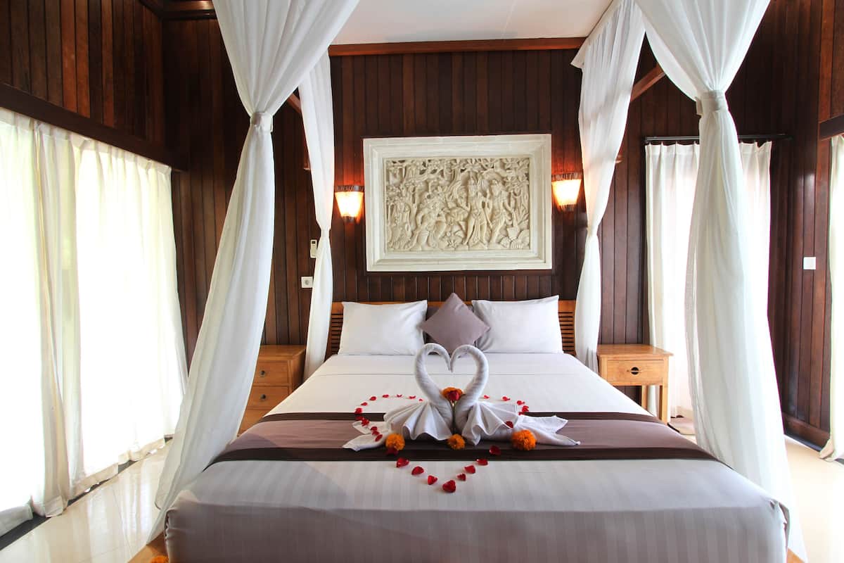 A hotel room with a large bed, white curtains, a wall-mounted light fixture, and a decorative wall piece.