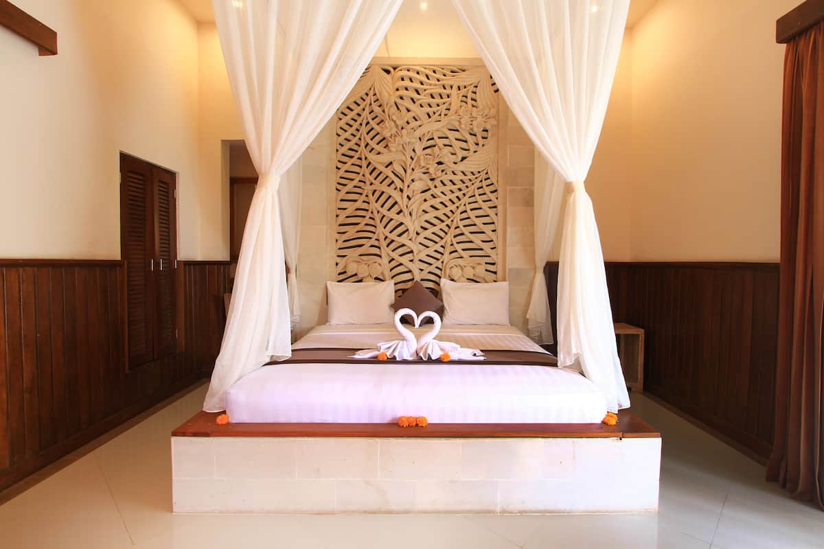 A four-poster canopy bed with a heart-shaped towel arrangement, a wooden headboard, and a decorative wall panel.