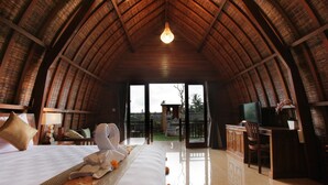 A spacious room with a high, curved wooden ceiling, a large bed with a neatly made bedspread, a sitting area with a chair and a table, and a television on a wooden cabinet. - Pondok Naya (Ubud)