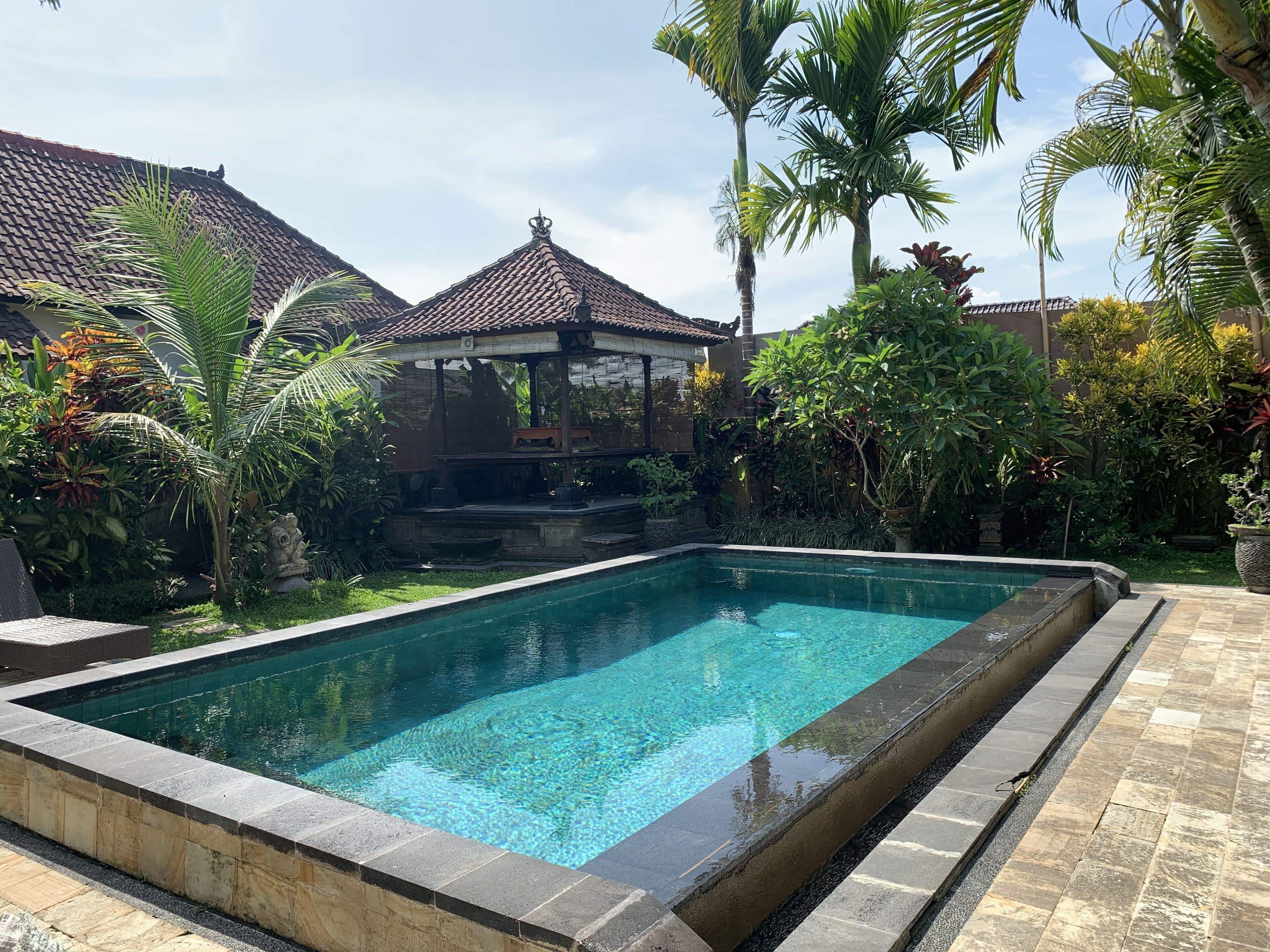 A rectangular swimming pool with clear water, surrounded by a stone deck. There is a gazebo with a tiled roof in the background, and various tropical plants around.