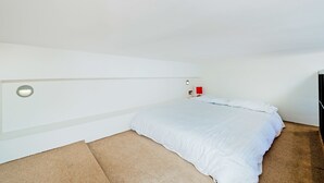 Une chambre à coucher avec un lit, une lampe fixée au mur et un objet rouge posé sur la table de chevet.