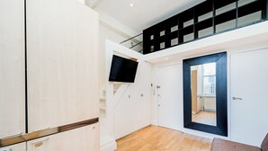 Un appartement moderne doté d’un plancher en bois, de meubles blancs, d’un téléviseur fixé au mur et d’un escalier menant à un niveau supérieur.