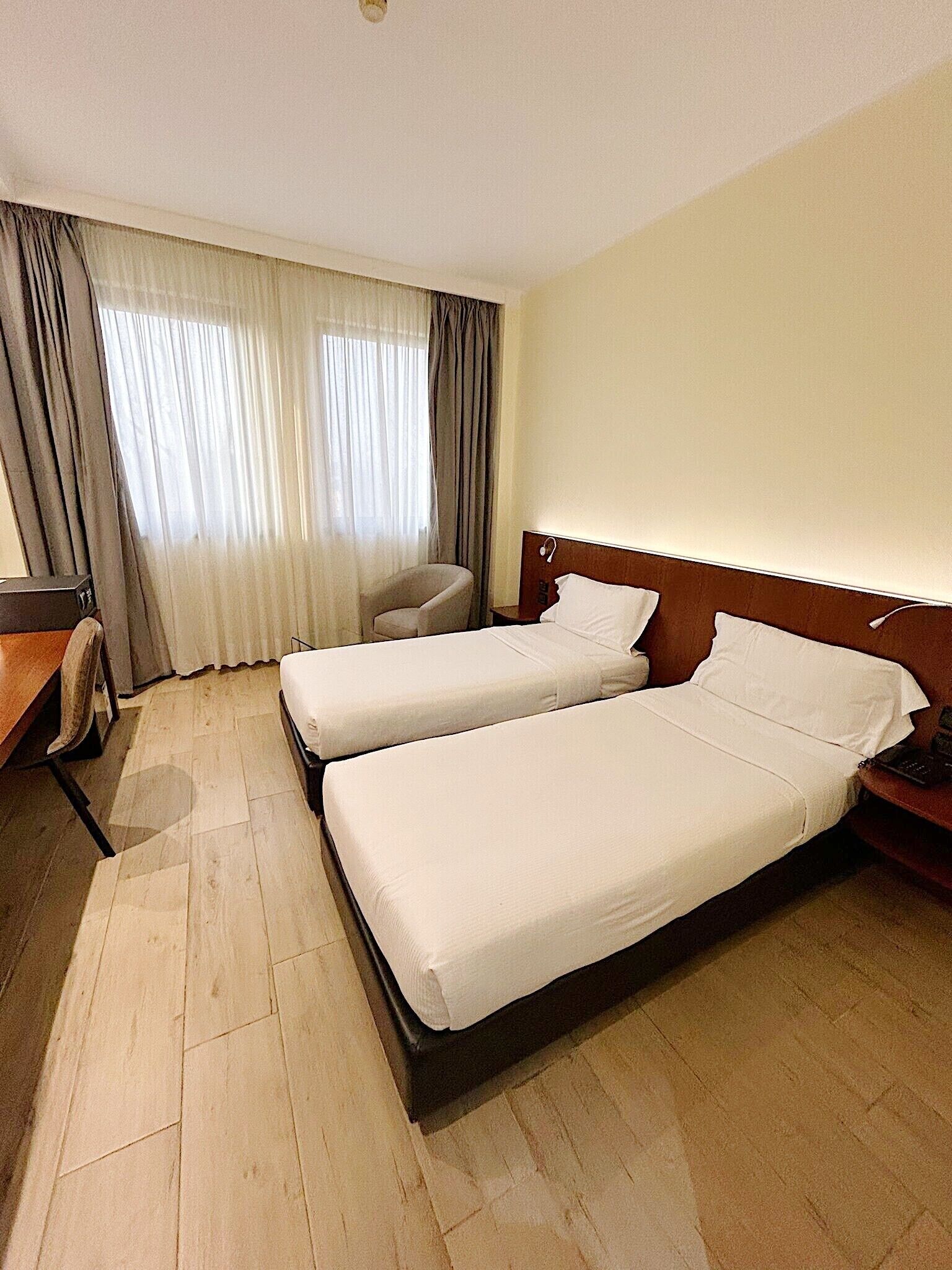 A hotel room with two beds, a desk, a chair, and a large window with curtains.