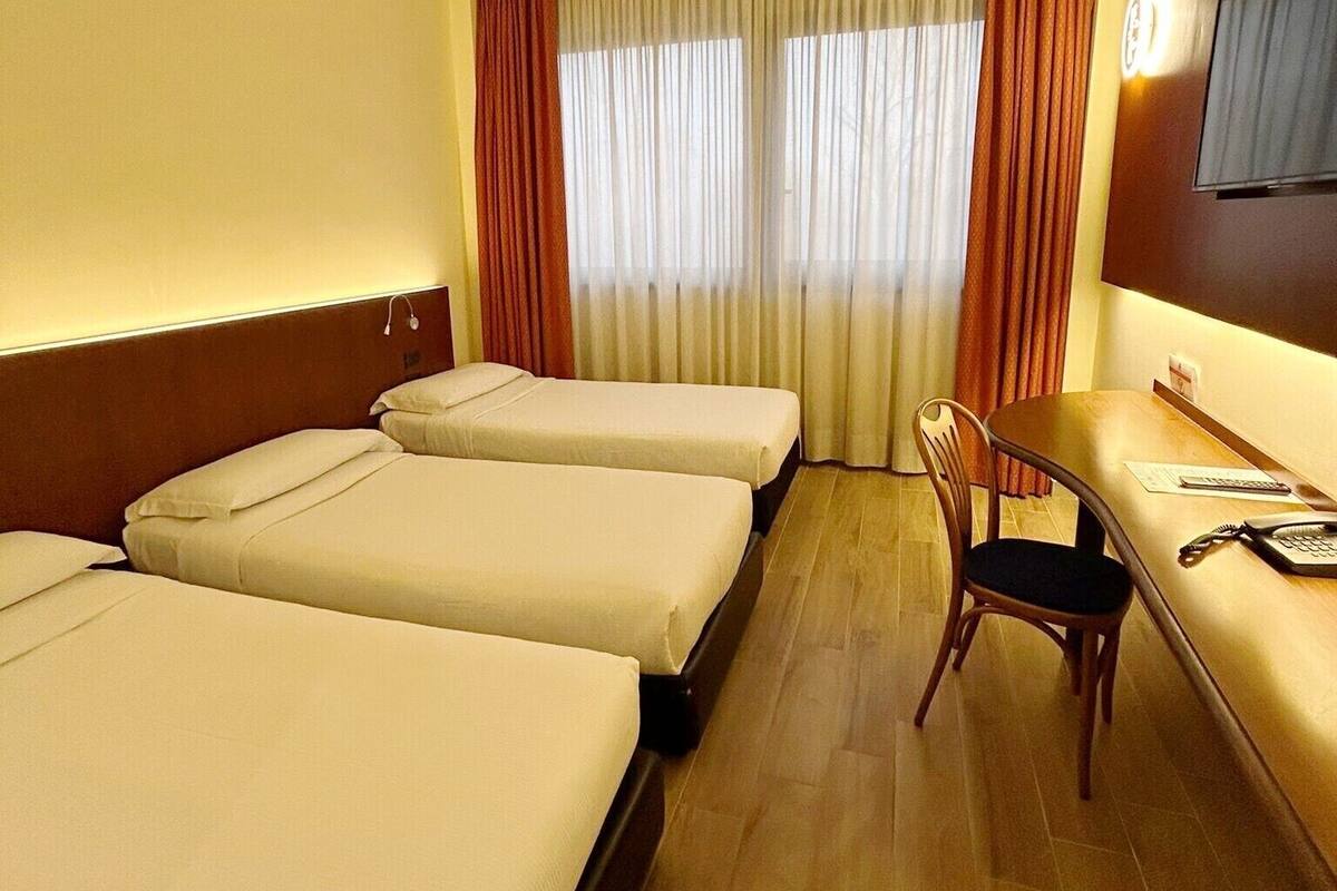 Une chambre d’hôtel avec deux lits simples, un bureau en bois avec une chaise, un téléviseur à écran plat fixé au mur et une fenêtre avec des rideaux.