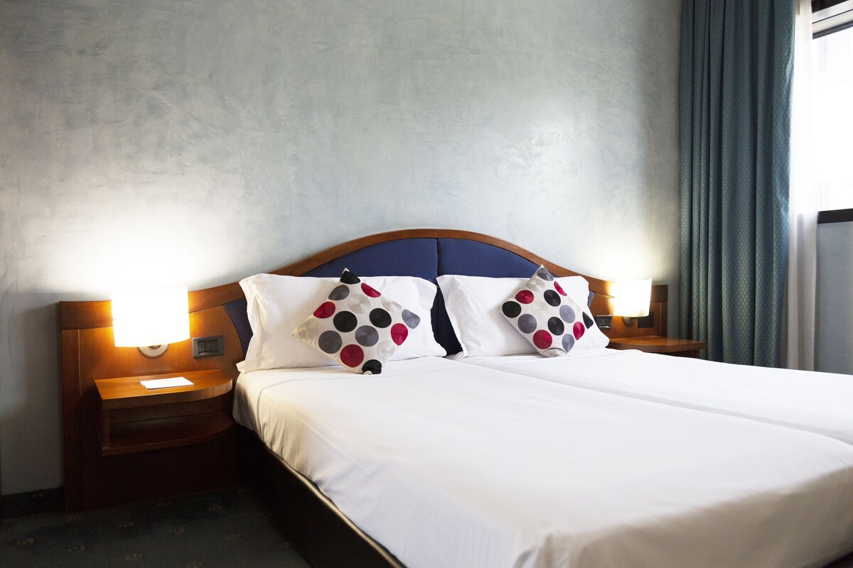 Habitación de hotel con una cama grande, dos mesitas de noche, un cabecero de madera y una ventana con cortinas.