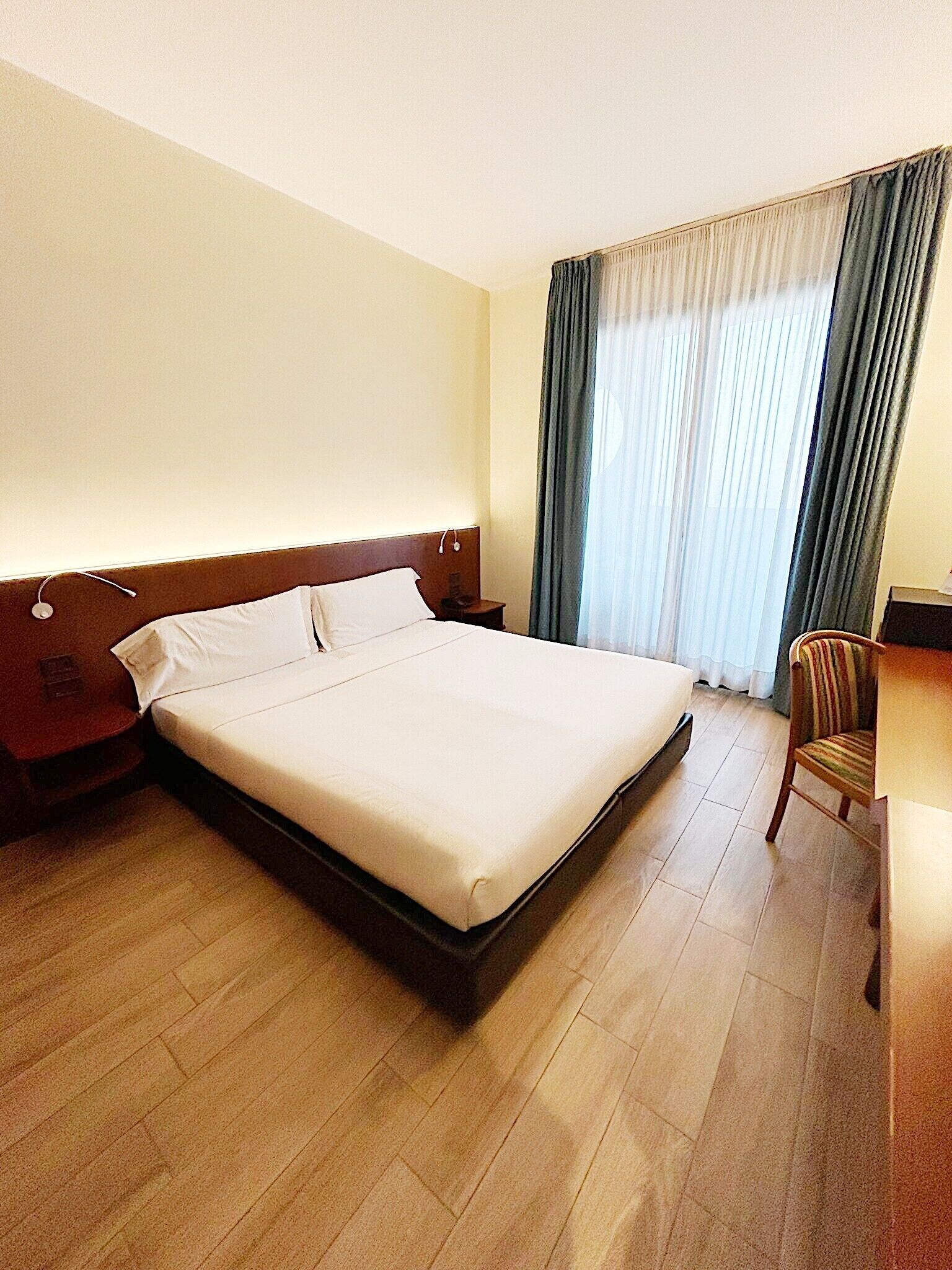 A hotel room with a large bed, a wooden headboard, a desk with a chair, and a window with curtains.