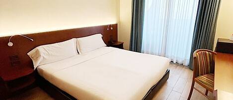 A hotel room with a large bed, a wooden headboard, a desk with a chair, and a window with curtains.