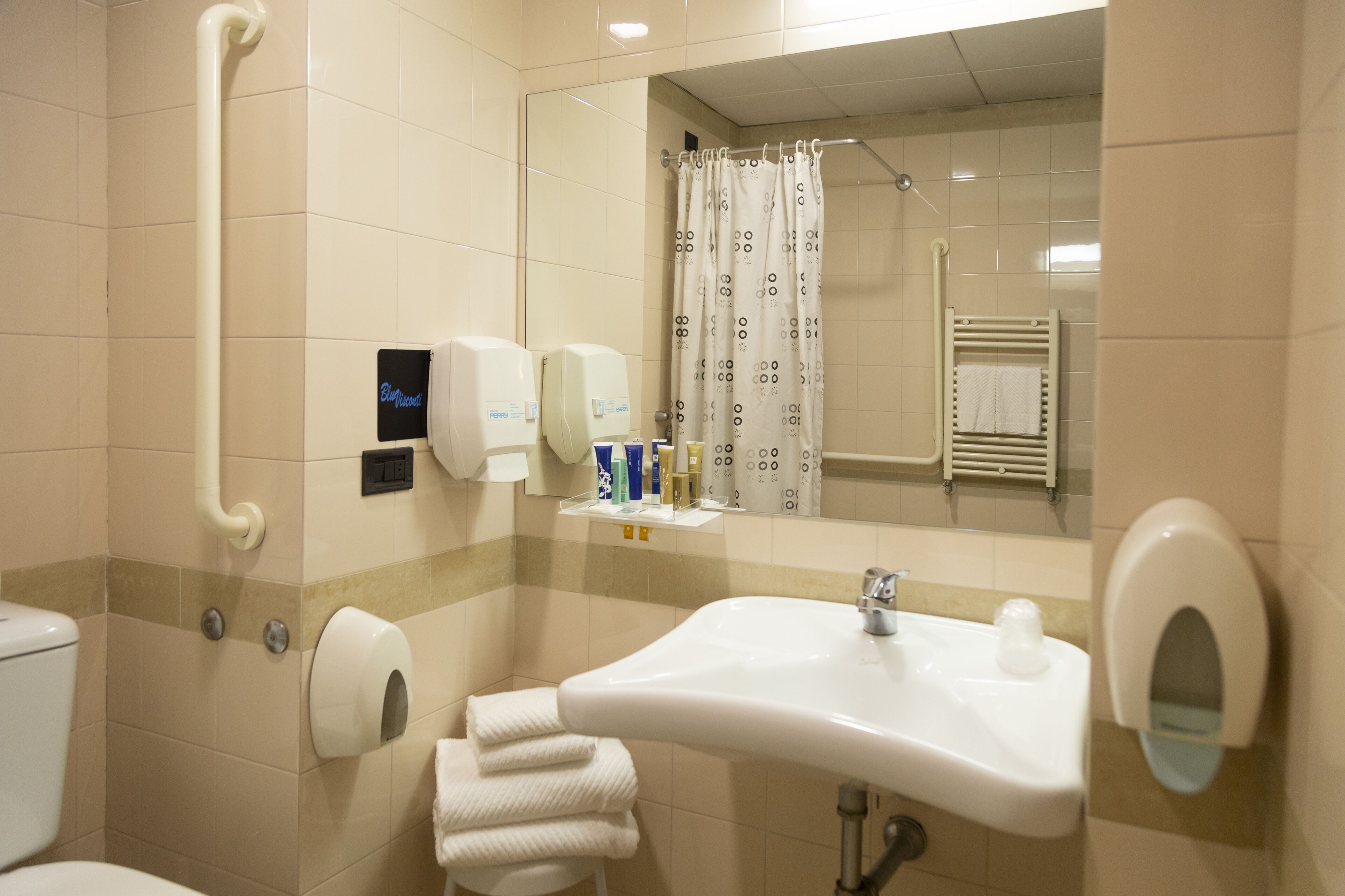 A bathroom with a sink, mirror, towel dispenser, and toilet.