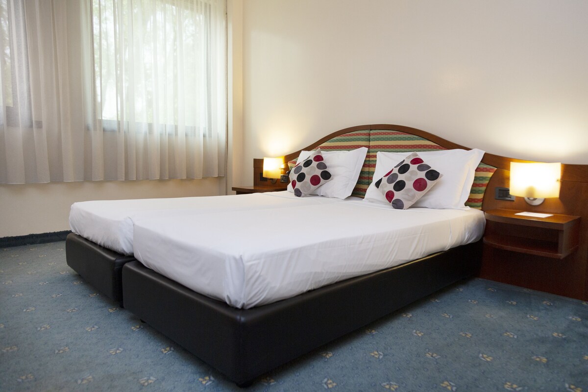 Una habitación de hotel con una cama grande, cabecera de madera, mesita de noche con lámpara y ventana con cortinas translúcidas.