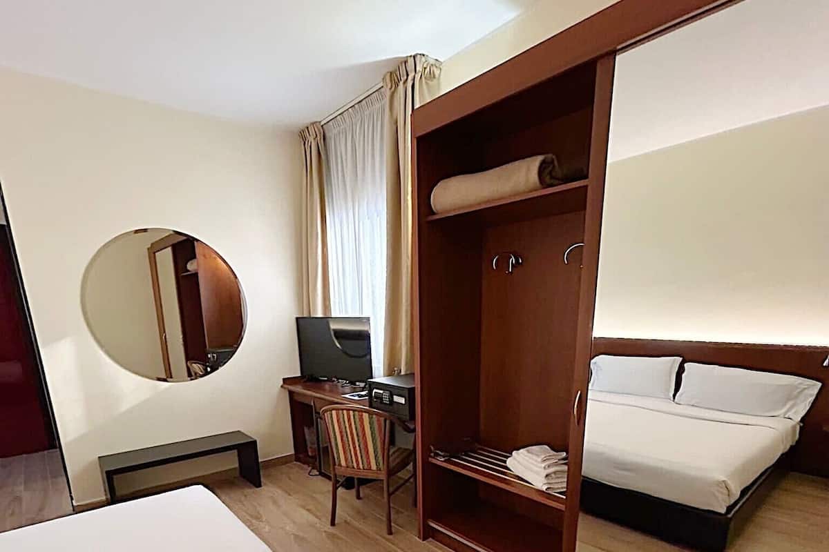 Une chambre d’hôtel avec un grand lit, un bureau avec un ordinateur, une chaise, une armoire et un miroir.