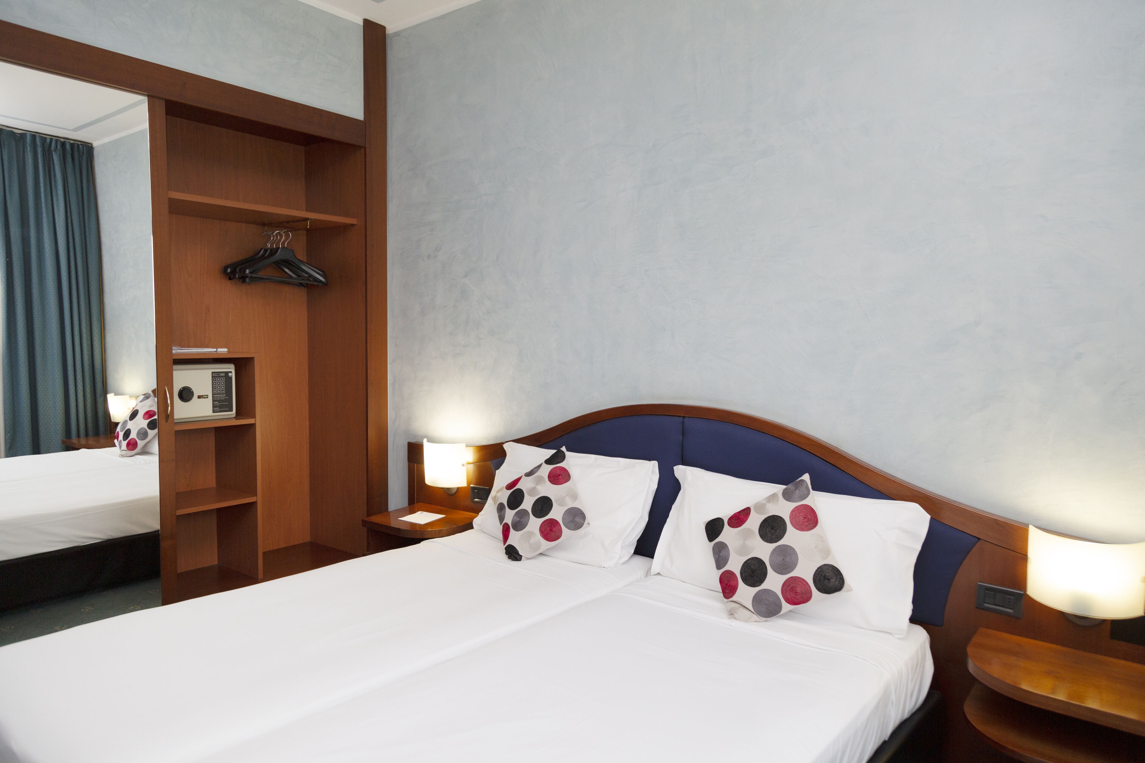 A hotel room with a large bed, a wooden wardrobe, a mirror, and a nightstand with a lamp.