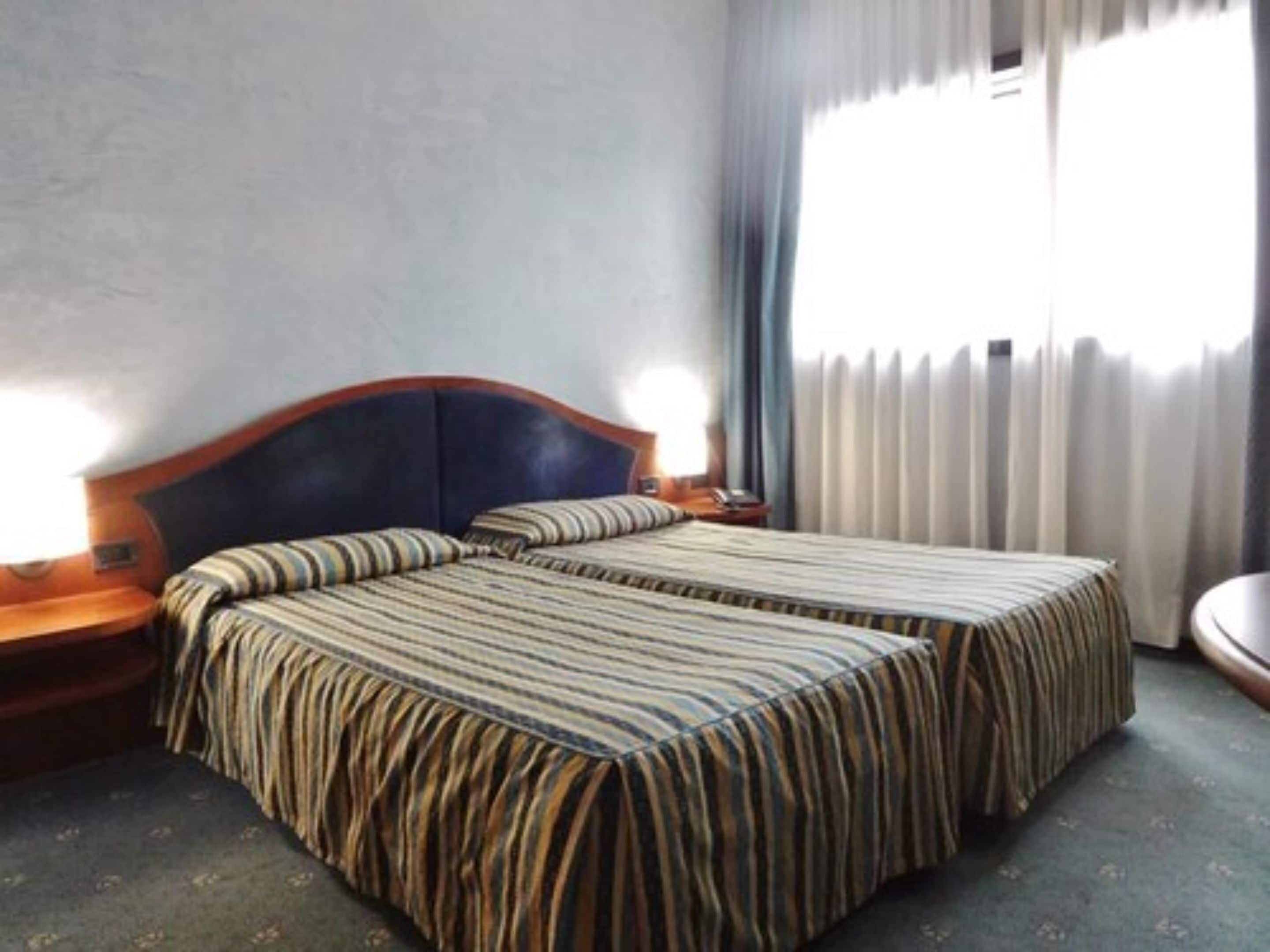 A double bed with striped bedding, a wooden headboard, bedside tables with lamps, and a window with curtains.