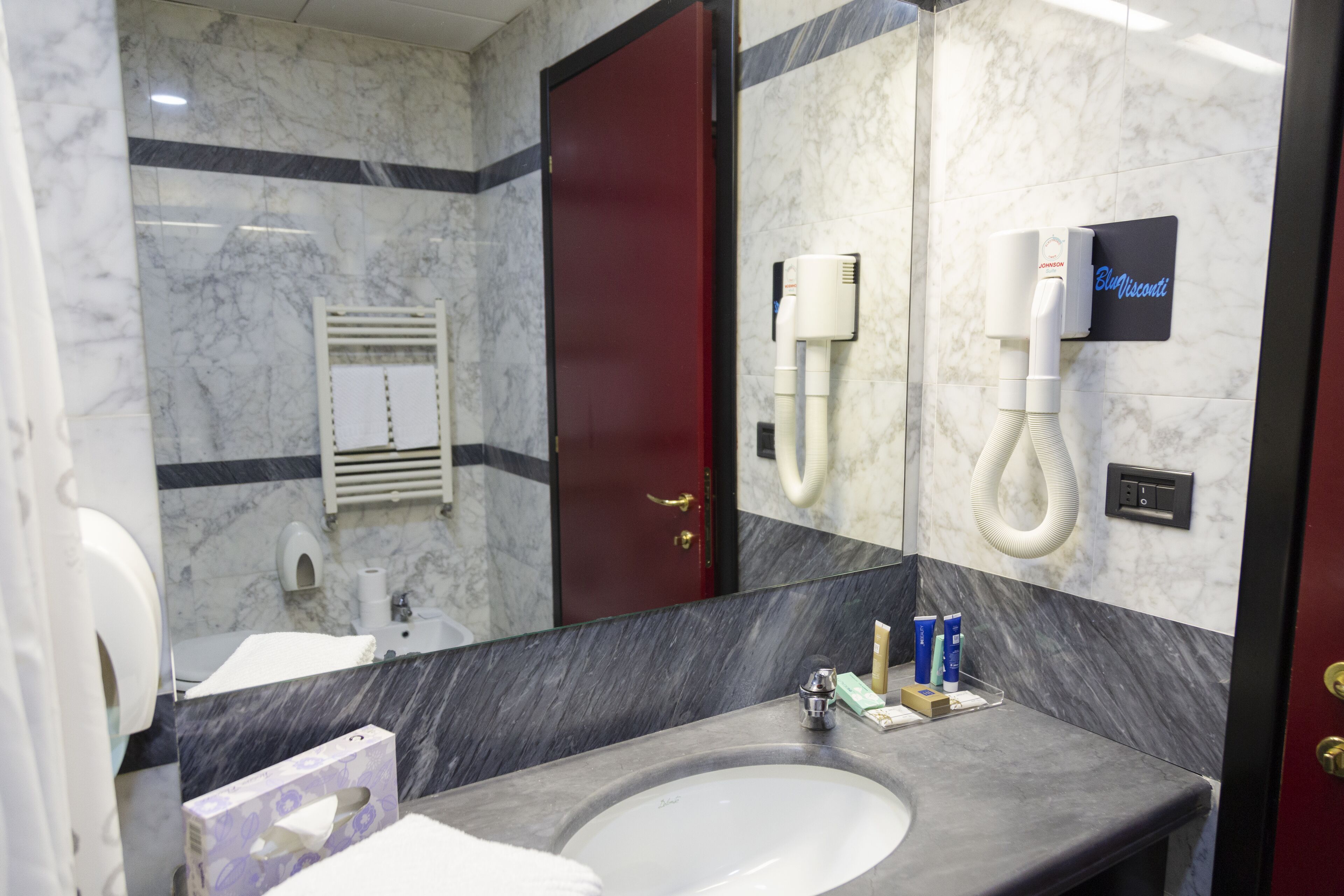 Un baño con un lavabo de mármol, una puerta roja, un espejo y un secador de manos.