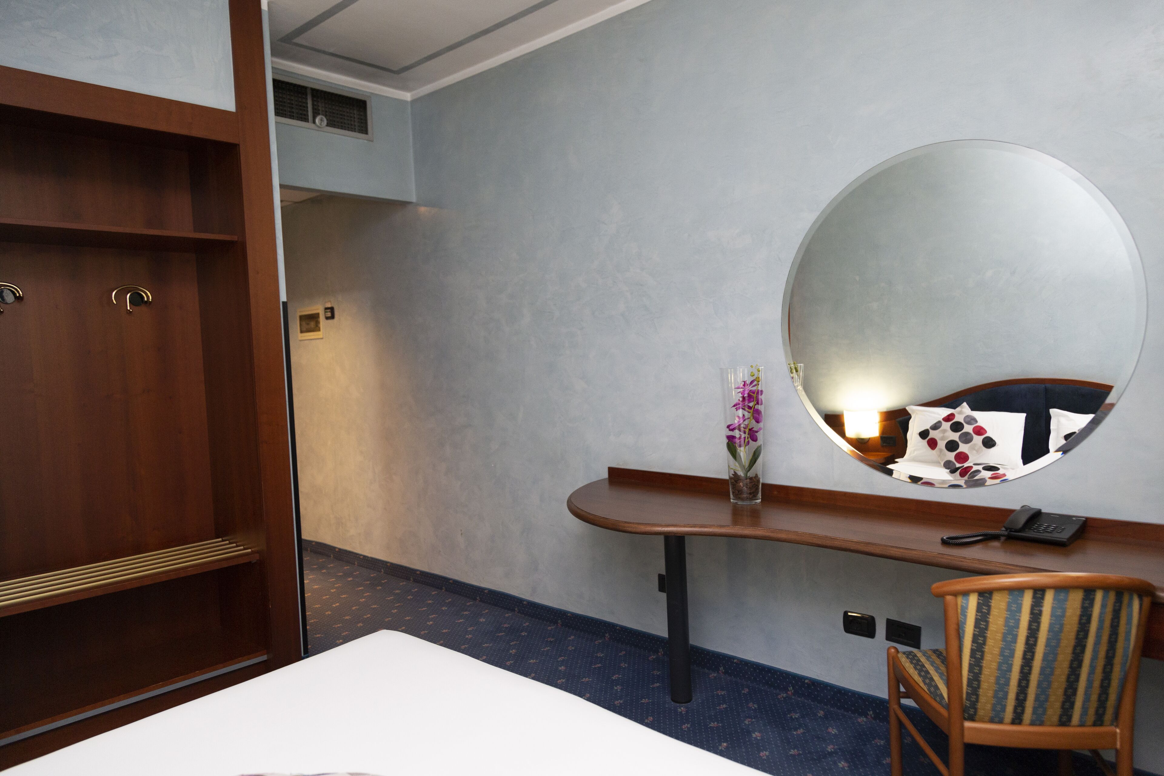 Quarto de hotel com cama, guarda-roupa de madeira, escrivaninha com espelho, cadeira e um vaso com flores.