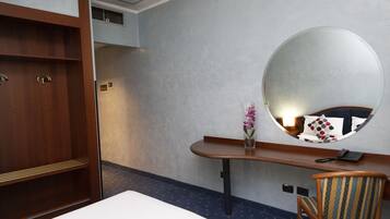 Quarto de hotel com cama, guarda-roupa de madeira, escrivaninha com espelho, cadeira e um vaso com flores.