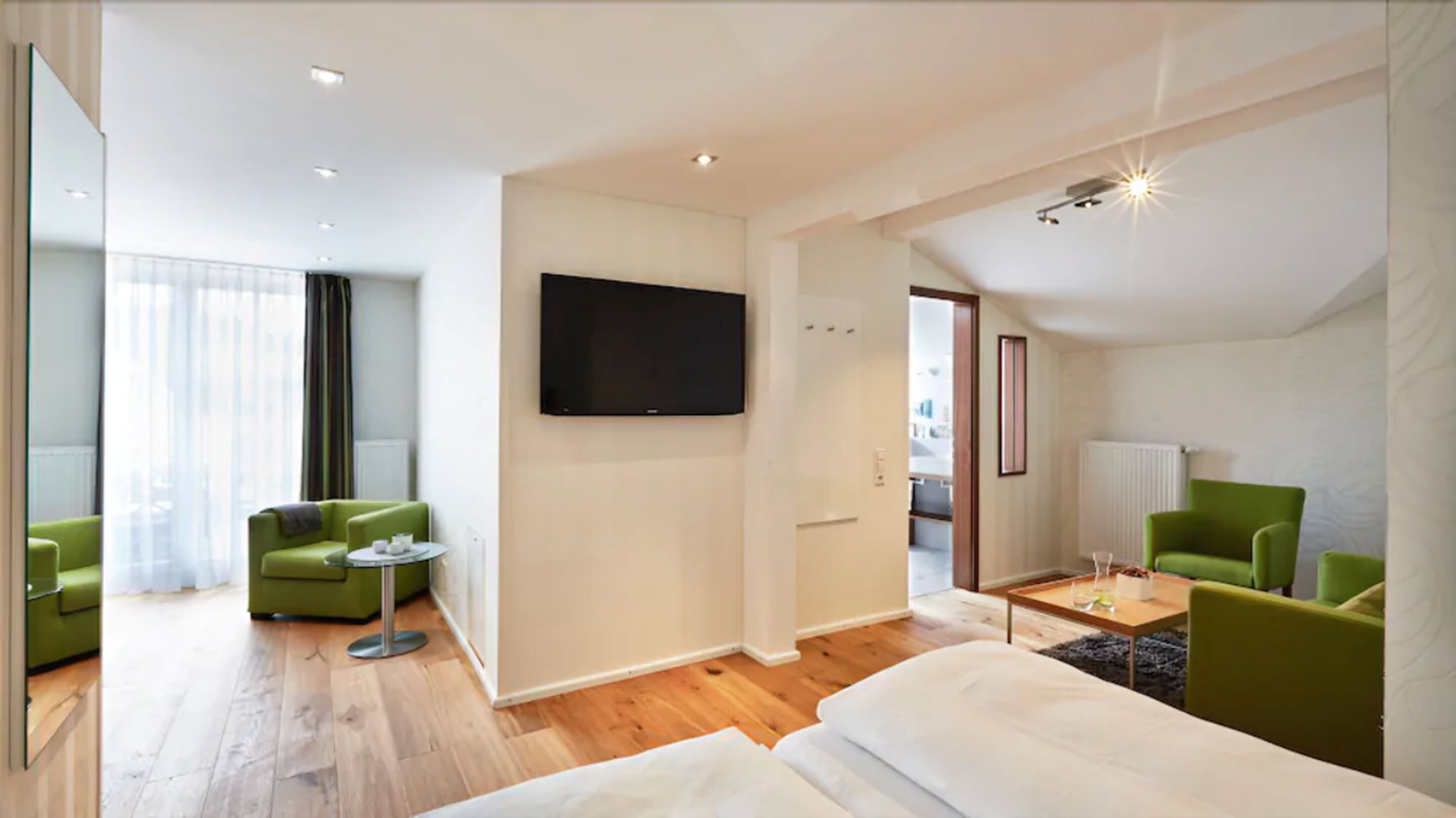 Una habitación de hotel moderna con una cama grande, un televisor de pantalla plana, sillones verdes y una pequeña mesa redonda.
