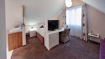 Een kleine hotelkamer met een bed, een bureau met televisie, een kitchenette en een raam met gordijnen.