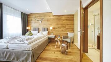 A modern hotel room with a wooden wall, a large bed, a chair, a small table with a lamp, and a shower area.