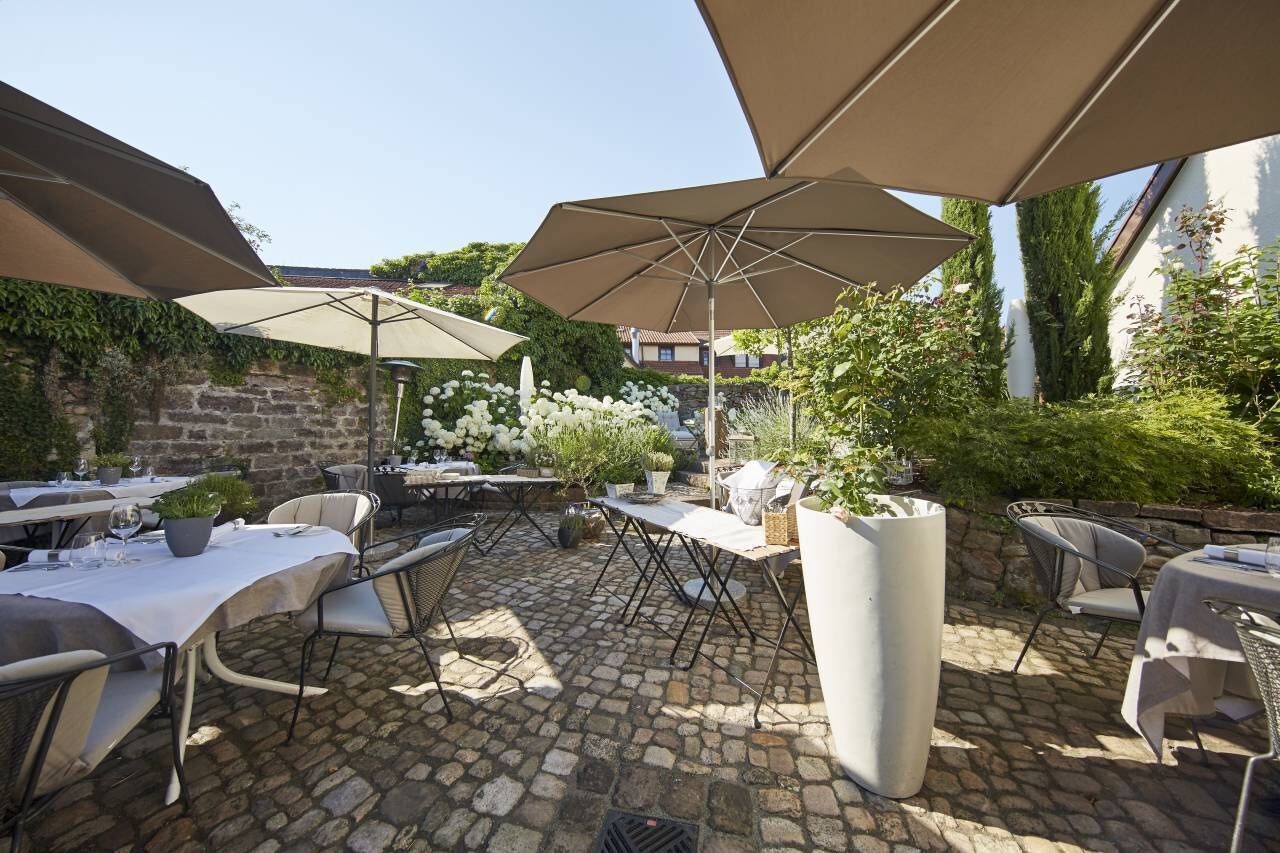 An outdoor patio area with stone flooring, tables with umbrellas, and potted plants.
