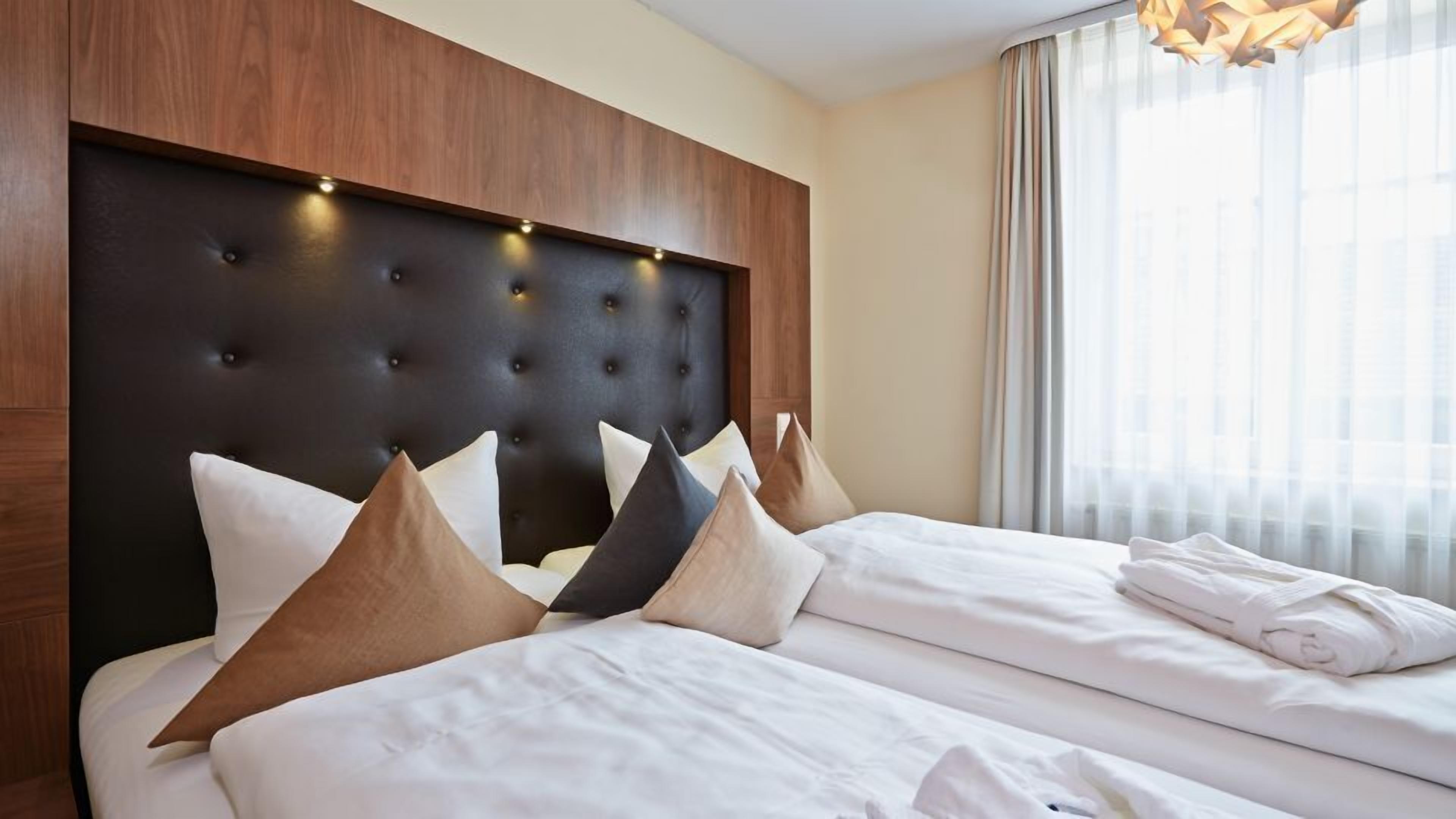 Habitación de hotel con una cama grande, varias almohadas, un cabecero de madera y una ventana con cortinas.