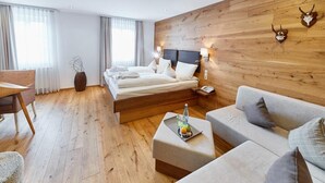 Habitación de hotel moderna con pared de madera, cama con ropa blanca, sofá gris, suelo de madera y una mesita con una botella y fruta.