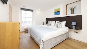 Ein modernes Schlafzimmer mit einem großen Bett, einem Nachttisch, einem an der Wand montierten Fernseher und einem Fenster mit Jalousien.