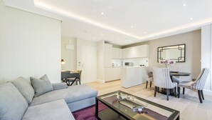 A modern living room with a grey sofa, a glass coffee table, and a purple rug. There is a dining area with striped chairs and a small kitchen in the background. - Imperial X (London)