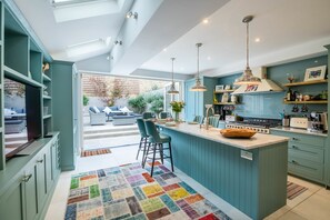 A modern kitchen with teal cabinetry, a central island, pendant lighting, and a view to a patio area. - Immaculate Designer Home in Wandsworth (London)