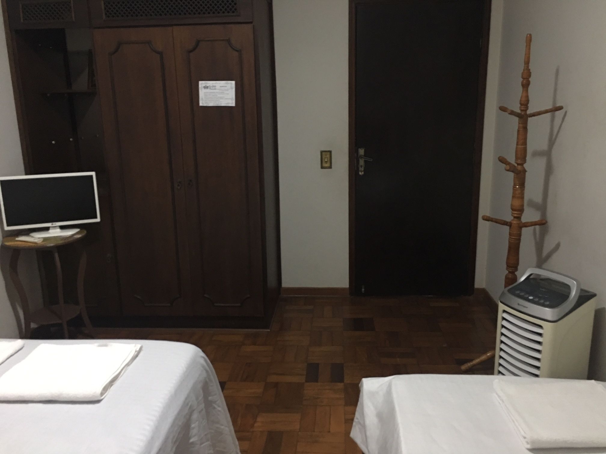 A hotel room with a wooden wardrobe, a desk with a computer, two beds with white linens, and a heater.