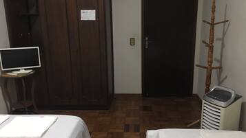 A hotel room with a wooden wardrobe, a desk with a computer, two beds with white linens, and a heater.