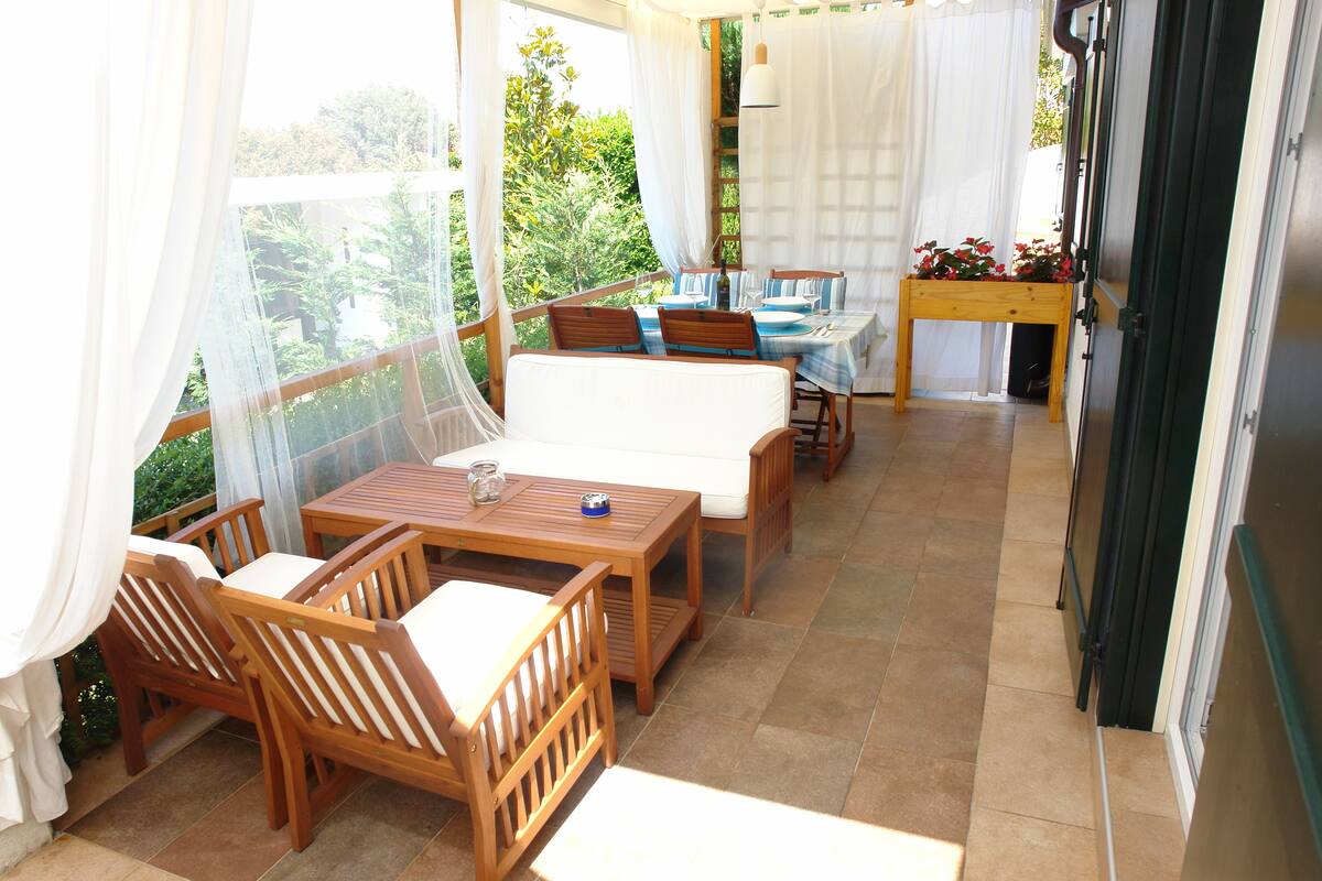A sunlit patio with wooden furniture, a small table with a blue tablecloth, and a view of greenery outside.