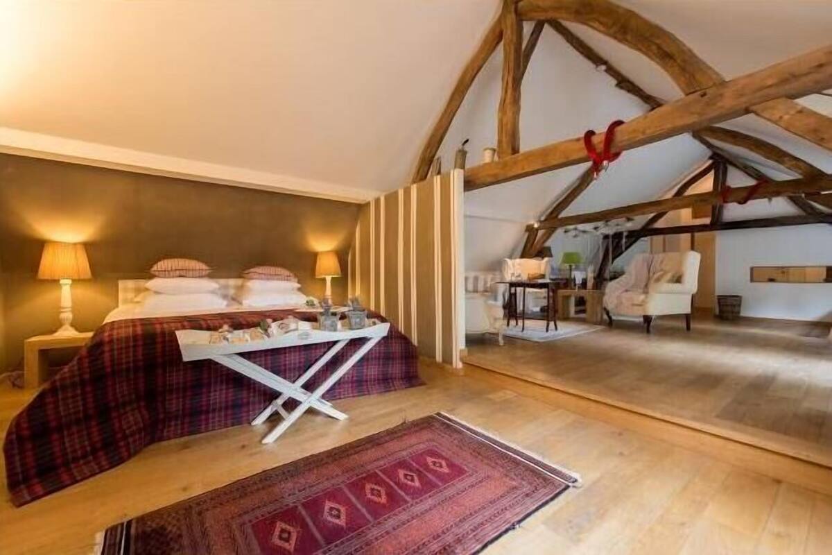 Ein gemütliches Schlafzimmer mit Bett, Nachttischlampen, einem kleinen Tisch und einem Sessel. Das Zimmer verfügt über Holzbalken an der Decke und einen Blick auf einen anderen Raum.