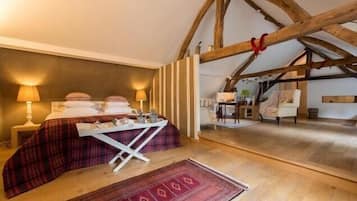 A cozy bedroom with a bed, bedside lamps, a small table, and a chair. The room has wooden beams on the ceiling and a view of another room.