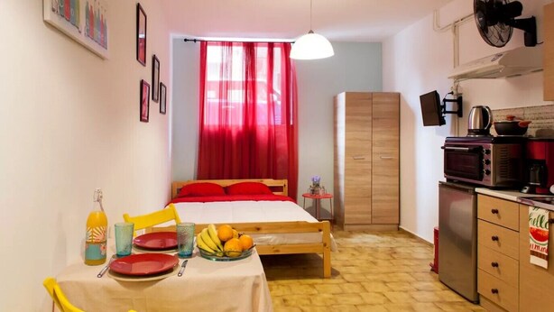 Une petite pièce comprenant un lit, une table à manger avec des chaises, un coin cuisine et un rideau rouge.