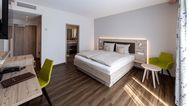 Habitación de hotel con piso de madera, una cama con ropa blanca, un escritorio con silla y una mesita con dos sillas.