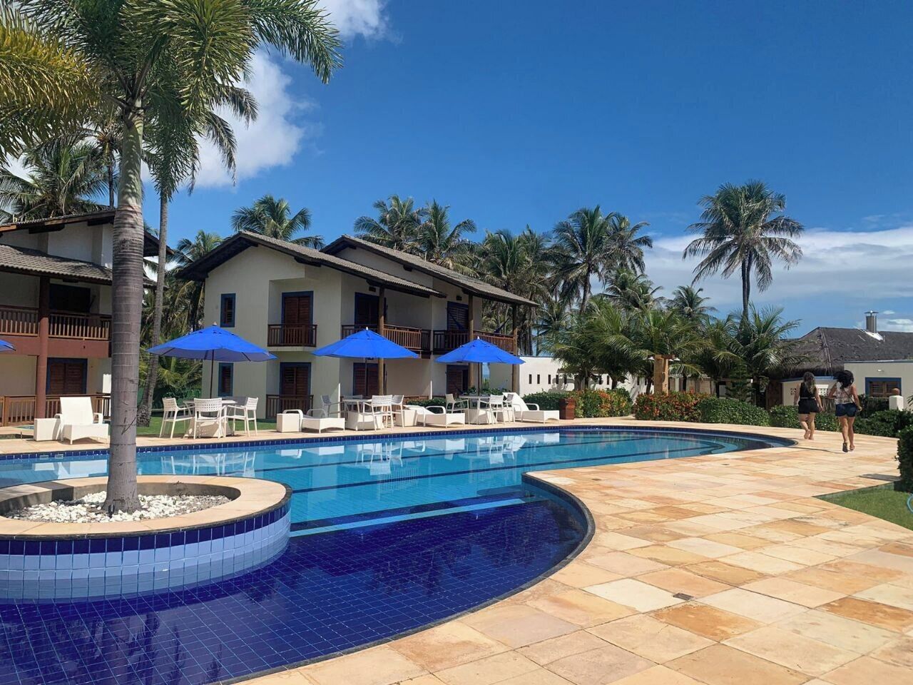 Una piscina con sillas de descanso y sombrillas, rodeada de palmeras y un edificio con balcones.