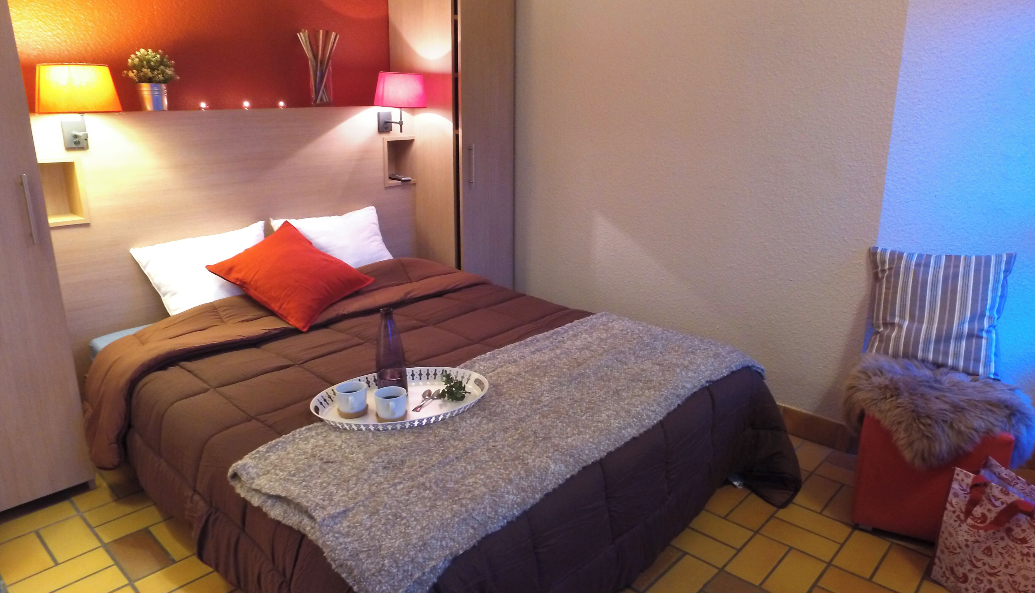 A hotel room with a bed, a tray with cups and a bottle, a red pillow, a lamp, and a small plant.