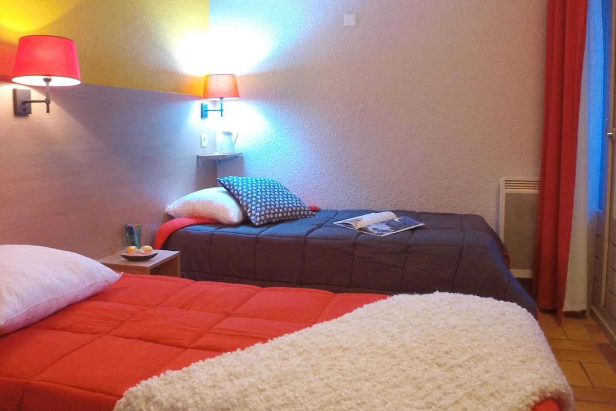A hotel room with two beds, each with a red and white blanket, a nightstand with a lamp, and a wall-mounted shelf with a book and a small plant.