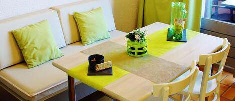 A dining area with a table, chairs, a sofa with cushions, a lamp, and a window with curtains.