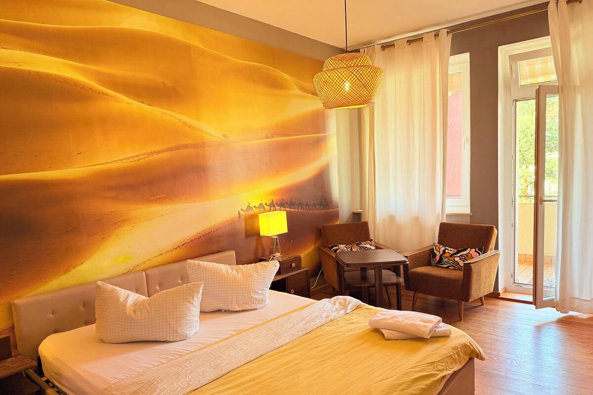 A hotel room with a large bed, a small table, two chairs, a lamp, and a view of the outdoors.
