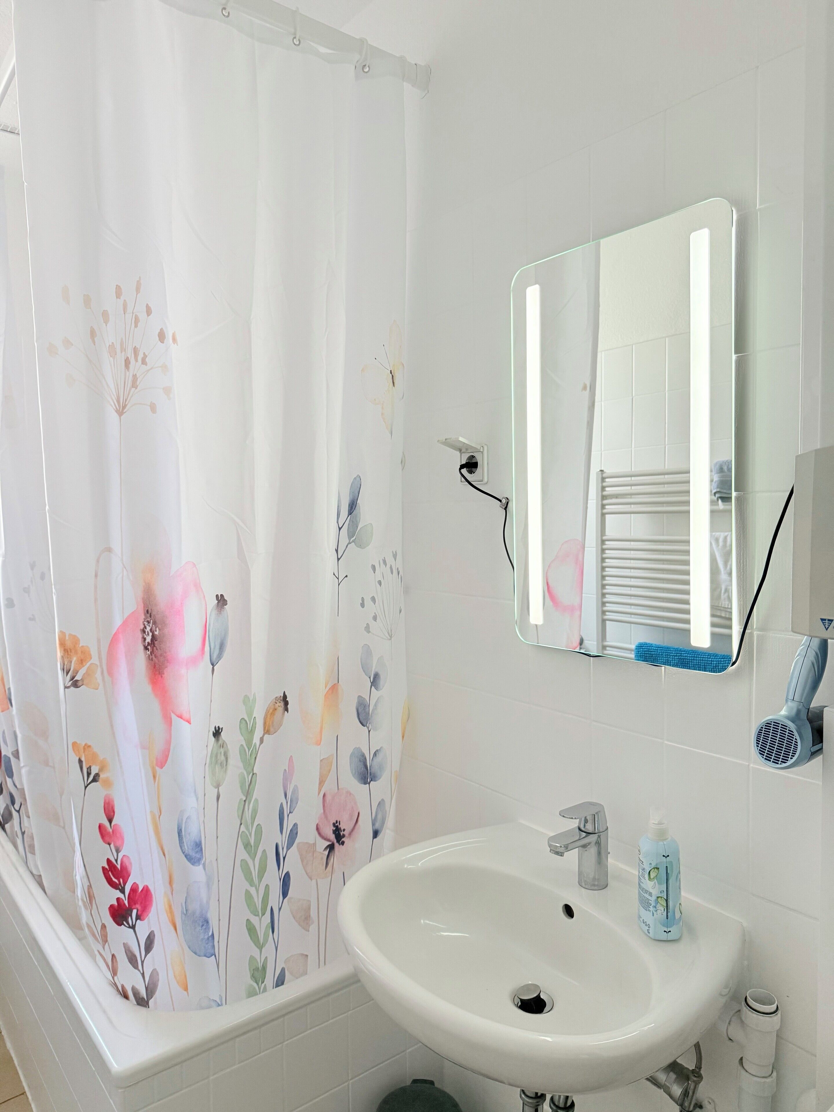 A bathroom with a white sink, a mirror, a shower curtain with floral patterns, and a hairdryer on the wall.