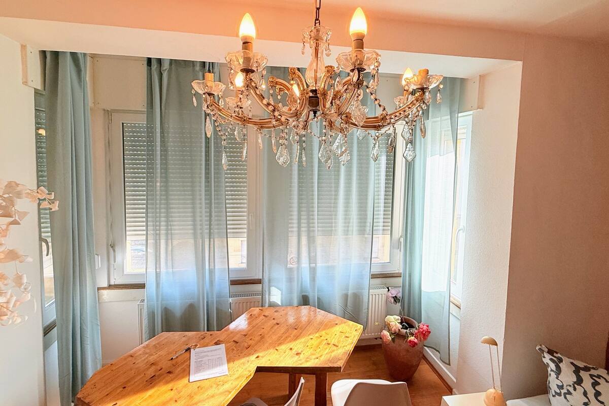A dining area with a wooden table, a chandelier, and a window with curtains.