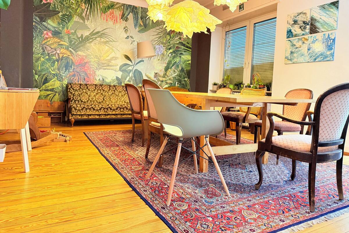 A dining area with a wooden table, chairs, a patterned rug, and a tropical mural on the wall.