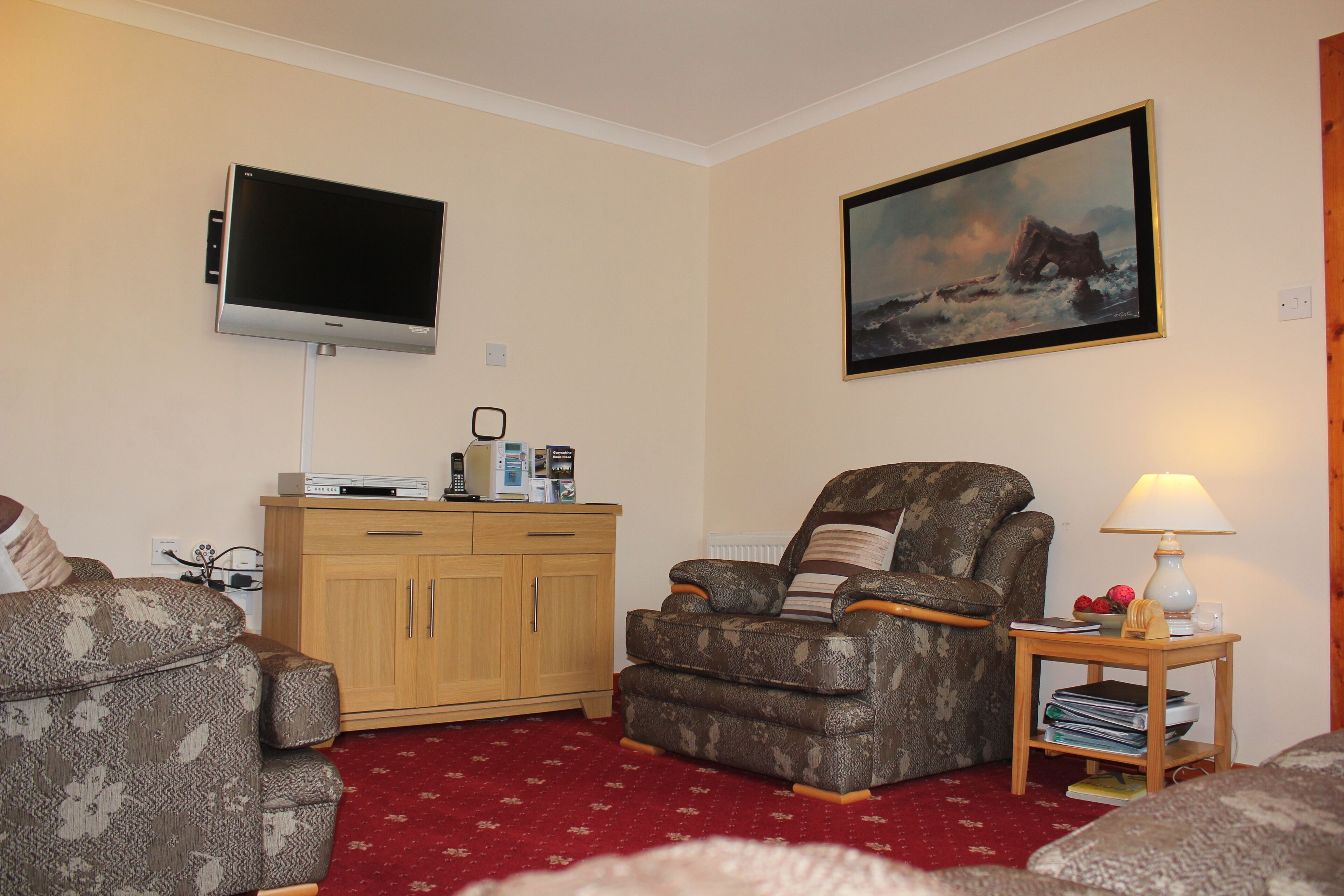 A living room with a flat-screen TV mounted on the wall, a wooden cabinet, a patterned armchair, a recliner, and a small table with a lamp and books.