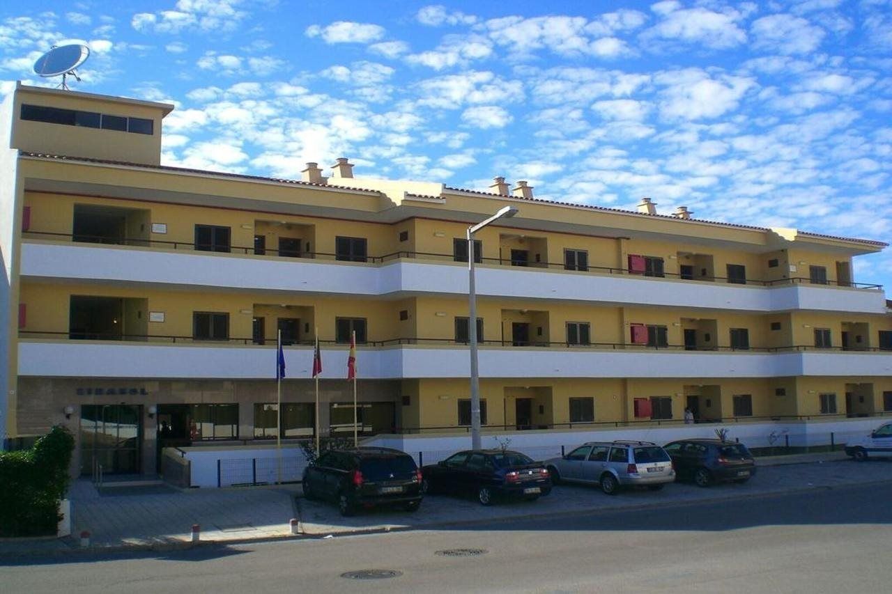 Un hotel de varios pisos con techo plano, entrada a nivel de calle y un estacionamiento con varios automóviles.
