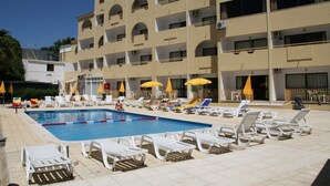 Una piscina con tumbonas y sombrillas, rodeada por un edificio de varios pisos con balcones.